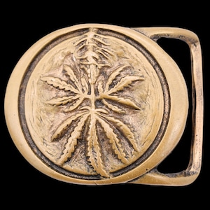 May include: A circular brass belt buckle featuring a raised, embossed design of a cannabis leaf centered within a smooth, rounded frame. The metal has a warm, golden-brown tone with darkened recessed areas that highlight the textured leaf details.