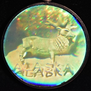 May include: A round, iridescent, silver-toned pendant with a raised image of a caribou and the word "ALASKA" in a stylized font.