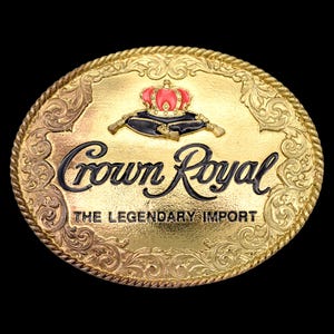 May include: Gold-toned oval belt buckle with the words "Crown Royal" in black script. The buckle features a red and gold crown design above the text and the words "THE LEGENDARY IMPORT" below. The border has a rope-like design.