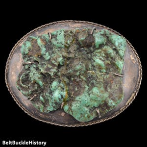 May include: An oval belt buckle featuring a large, raw, textured turquoise stone with green and brown mineral inclusions, secured by metal prongs onto a dark, tarnished silver-toned base with a decorative twisted rope-style border edge.