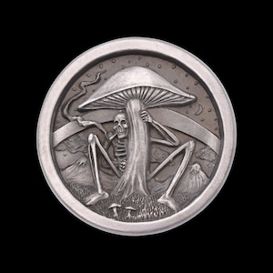 May include: A silver-colored round metal object with a detailed relief design. The design features a skeleton seated under a large mushroom, with mountains and a crescent moon in the background. The object has a dark background.
