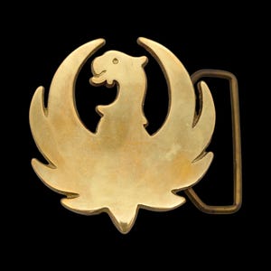 K&ouml;nnte beinhalten: Goldschnalle mit stilisiertem Ph&ouml;nix-Design. Die Schnalle hat einen rechteckigen Rahmen und eine detaillierte Darstellung des mythischen Vogels mit ausgebreiteten Fl&uuml;geln. Der Hintergrund ist einfarbig schwarz.