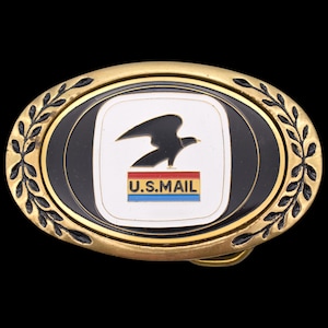 May include: Oval-shaped belt buckle with a gold-colored frame and black and white center. The center features the U.S. Mail logo: a black eagle over a red, white, and blue bar, with the text "U.S. MAIL" below. The buckle has a decorative leaf design.