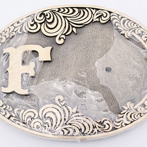 May include: Oval-shaped metal belt buckle with an ornate design. The buckle features a large letter "F" in the center, surrounded by a detailed floral pattern. The color scheme is a mix of gold and silver, giving it a vintage look.
