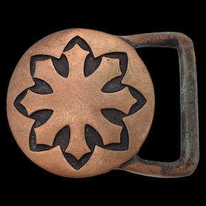 May include: A copper-colored belt buckle with a circular design featuring a stylized eight-pointed star. The buckle has a rectangular bar for the belt and shows signs of wear, giving it a vintage appearance.