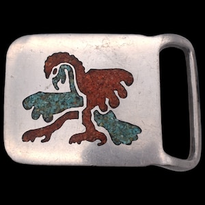 May include: A rectangular silver-toned belt buckle with an abstract design. The design features a bird-like figure and other shapes, inlaid with turquoise and red-orange stones. The buckle has a simple, sturdy design.