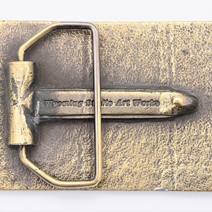 Puede incluir: Hebilla de cintur&oacute;n rectangular de color bronce con una superficie texturizada. La hebilla presenta un marco rectangular y una barra central con el texto "Wyoming State Art Works".