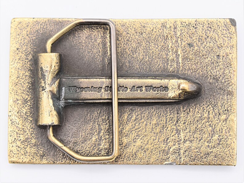 Puede incluir: Hebilla de cintur&oacute;n rectangular de color bronce con una superficie texturizada. La hebilla presenta un marco rectangular y una barra central con el texto "Wyoming State Art Works".