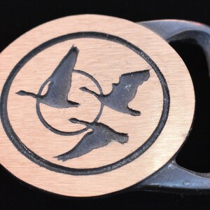 May include: A vintage metal belt buckle with a circular design featuring three flying geese within a circle. The background is a light brown color, and the geese are dark gray. The buckle has a silver-colored frame.
