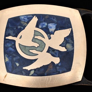 May include: A rectangular copper-colored belt buckle with a dark blue and white stone inlay. The inlay features a white silhouette of two birds in flight. The buckle has a dark brown loop.