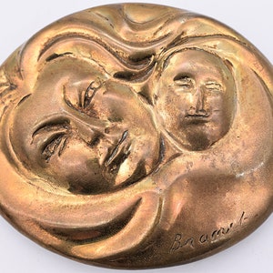May include: A bronze-toned, circular relief sculpture featuring two faces. One face is larger, with closed eyes and flowing hair, embracing a smaller face. The artist's signature, "Bramit," is visible.