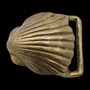 May include: A brass-colored belt buckle with a seashell design. The buckle features a detailed, textured surface with radiating lines, and a rectangular loop for the belt strap. The background is black.
