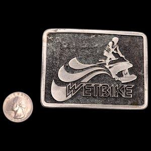 May include: A rectangular silver-toned belt buckle with a textured surface. The buckle features a stylized image of a person riding a jet ski, with the word "WETBIKE" below. A U.S. quarter is next to the buckle for size comparison.