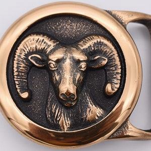 May include: A bronze-colored belt buckle featuring a detailed ram's head within a circular frame. The ram's head is centrally positioned against a dark background, with the buckle's outer edges in a lighter bronze hue. The buckle has a rectangular shape with a curved edge.