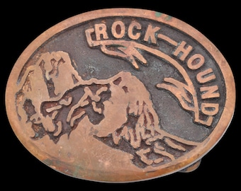 May include: Oval copper belt buckle with the words "ROCK-HOUND" arched across the top. The buckle features a mountain range design in the centre. The buckle has a dark patina and is set against a black background.