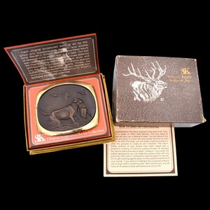 May include: A bronze sculpture of a dog retrieving a duck, mounted on a square, gold-colored plaque. The plaque is in a red and gold box with the text "SK Original Sculptured Buckle Collection".