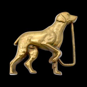 May include: A gold-colored metal belt buckle in the shape of a dog, standing with its head up and a rectangular bar attached. The dog is detailed with visible muscles and a smooth finish. The buckle is set against a black background.