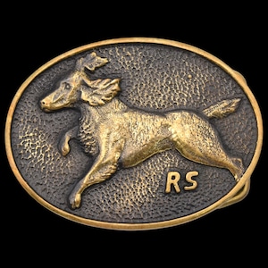 May include: Oval-shaped brass belt buckle featuring a detailed relief of a running dog. The dog is in a dynamic pose, with the initials "RS" below. The buckle has a textured background and a dark, aged finish.