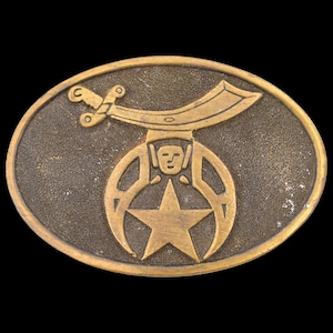 May include: Oval-shaped, antique brass belt buckle. The buckle features a central star, a stylized face, and a curved sword design. The buckle has a weathered appearance, suggesting age and use.