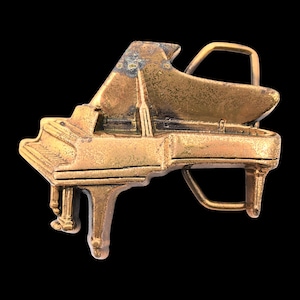 May include: A vintage brass-colored belt buckle in the shape of a grand piano. The buckle has a detailed design with visible keys, legs, and a raised lid. The metal has a slightly aged appearance, adding to its vintage charm.