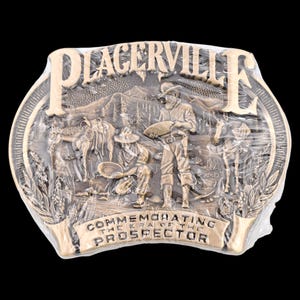 May include: A bronze-colored belt buckle with the word "PLACERVILLE" at the top. The buckle features a detailed relief scene of prospectors, horses, and mountains. The text "COMMEMORATING THE ERA OF THE PROSPECTOR" is at the bottom.