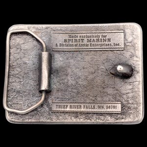 May include: A rectangular silver-colored belt buckle with a textured surface. The buckle has a metal loop and a small, rounded design element. The text "Made exclusively for SPIRIT MARINE" and "THIEF RIVER FALLS, MN, 56701" are visible.