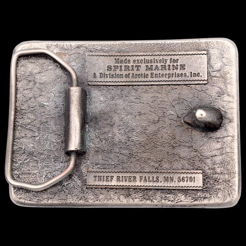 May include: A rectangular silver-colored belt buckle with a textured surface. The buckle has a metal loop and a small, rounded design element. The text "Made exclusively for SPIRIT MARINE" and "THIEF RIVER FALLS, MN, 56701" are visible.