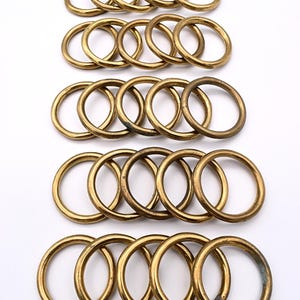 May include: A collection of brass-colored metal rings arranged in rows. Each ring is circular with a consistent thickness. The rings have a slightly aged appearance, suggesting they may be vintage or have a rustic finish. The background is plain white.