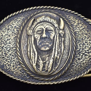 May include: Oval-shaped brass belt buckle featuring a detailed relief of a Native American chief in a headdress. The buckle has a textured background and a rope-like border, with a brass clasp.