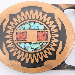 May include: A copper-colored belt buckle with a circular design. The center features a black outline with turquoise and red stone inlays. The buckle has a rectangular frame and a hole for attachment.