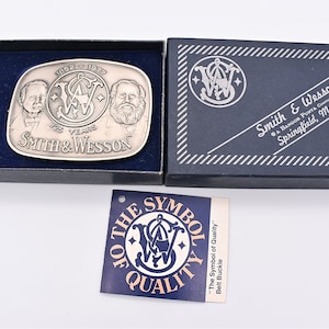May include: A silver belt buckle with a Smith & Wesson logo and the text "1852-1972 125 YEARS SMITH & WESSON". The buckle is in a blue box with a white label that says "THE SYMBOL OF QUALITY" and "The Symbol of Quality" Belt Buckle.