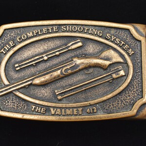 May include: A rectangular, bronze-colored belt buckle with the text "THE COMPLETE SHOOTING SYSTEM" and "THE VALMET 412". The buckle features an oval design with a rifle and other gun parts.