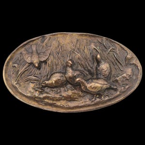 May include: An oval, bronze-colored relief sculpture depicting a marsh scene with birds. The detailed artwork shows several birds, including some in flight, amidst tall reeds and grasses. The overall design has a vintage aesthetic.