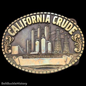 May include: An oval brass belt buckle featuring a relief design of an oil refinery with tall storage tanks and two oil derricks. The text CALIFORNIA CRUDE is embossed along the top edge, and a blank banner is positioned at the bottom center.