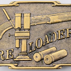 May include: A rectangular, bronze-colored belt buckle with a detailed design. The buckle features a rifle, reloading equipment, and the word "RELOADER". Two bullet casings are also visible. The buckle has a textured background.
