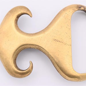 May include: A vintage-style brass belt buckle with a unique, flowing design. The buckle has a curved shape with an open loop on one end and a hook-like element on the other. The brass has a slightly aged appearance.