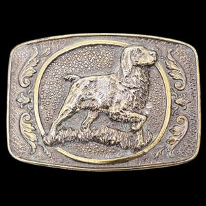 May include: Rectangular metal belt buckle with a detailed relief of a dog within a circular frame. The dog is depicted in a running pose, surrounded by textured details and decorative flourishes. The buckle has a two-tone finish.