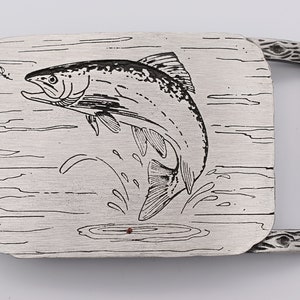 May include: Silver-toned metal belt buckle featuring a leaping fish design. The fish is depicted in black line art, with water and a fly also illustrated. The buckle has a textured, wood-grain style frame.