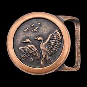 May include: A copper-toned metal belt buckle with a circular design. The buckle features a detailed relief of two ducks with outstretched wings and several flying birds against a dark background. The buckle has a rectangular bar for the belt.