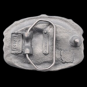 May include: A silver-colored metal belt buckle with a textured surface. The back of the buckle features a serial number plate, the text "COPYRIGHT 1984", and a small emblem. The buckle has a metal loop for the belt.