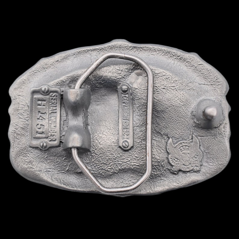 May include: A silver-colored metal belt buckle with a textured surface. The back of the buckle features a serial number plate, the text "COPYRIGHT 1984", and a small emblem. The buckle has a metal loop for the belt.