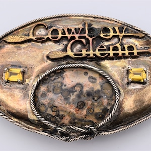 May include: Oval-shaped belt buckle with the words "Cowboy Heaven" in gold lettering. The buckle has a silver and bronze finish, two yellow gemstones, and a rope-like border. The center has a speckled pattern.