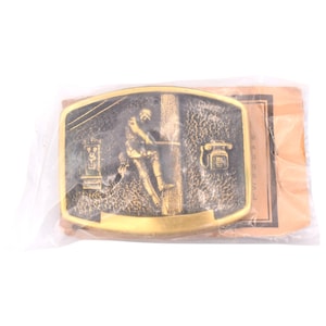 May include: A rectangular, gold-colored belt buckle featuring a relief of a telephone lineman working on a utility pole. The buckle has a vintage aesthetic and is packaged in a clear plastic bag with a brown paper backing.