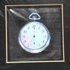 May include: A silver pocket watch with a white face and black Roman numeral hour markers. The watch has a small second hand dial at the bottom. The watch is framed in a black and gold frame.