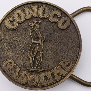 May include: A vintage brass belt buckle with the word "CONOCO" at the top and "GASOLINE" at the bottom. The buckle features a colonial-era figure in the center. The buckle has a rectangular loop for the belt.