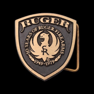 May include: A vintage brass belt buckle with a shield shape. The buckle features the word "RUGER" at the top, with "30 YEARS OF RUGER FIREARMS" around a central emblem. The years "1949-1979" are also displayed.