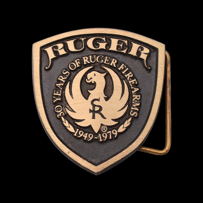 May include: A vintage brass belt buckle with a shield shape. The buckle features the word "RUGER" at the top, with "30 YEARS OF RUGER FIREARMS" around a central emblem. The years "1949-1979" are also displayed.