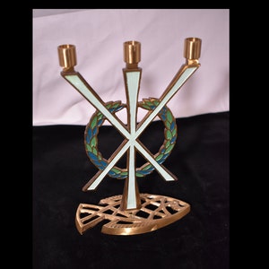 May include: A brass three-armed candelabra with a white and green enamel design. The design features a stylized cross with a wreath around it. The base of the candelabra is shaped like a fish.