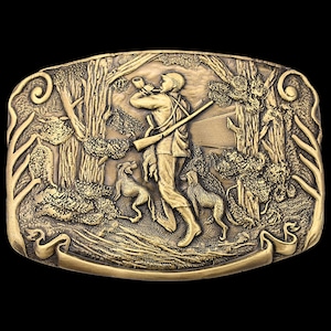 May include: A gold-colored metal belt buckle with a detailed relief scene of a hunter in a forest with two dogs. The hunter is blowing a horn and carrying a rifle. The buckle has a rectangular shape with decorative scrollwork.