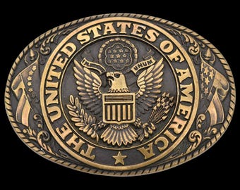 May include: Oval-shaped belt buckle with a detailed relief of the United States of America's Great Seal. The design features an eagle, stars, and the text "UNITED STATES OF AMERICA". The buckle has a bronze and black finish.